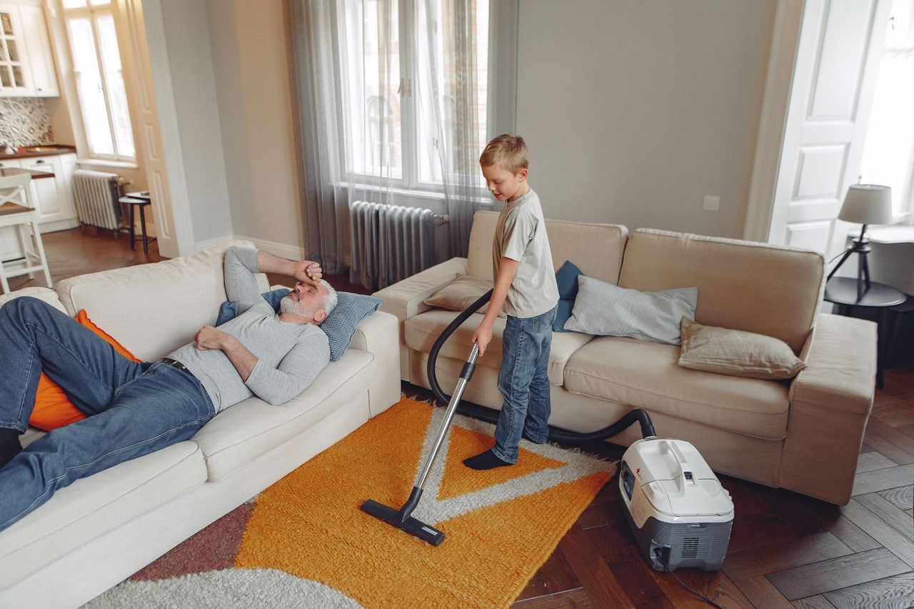 A young boy cleans the living room carpet with a vacuum while his grandfather rests on the sofa.